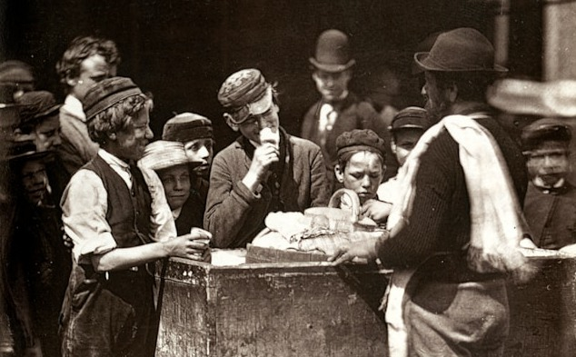 A vintage black and white photograph of people gathered around a cart in an old-world street scene.