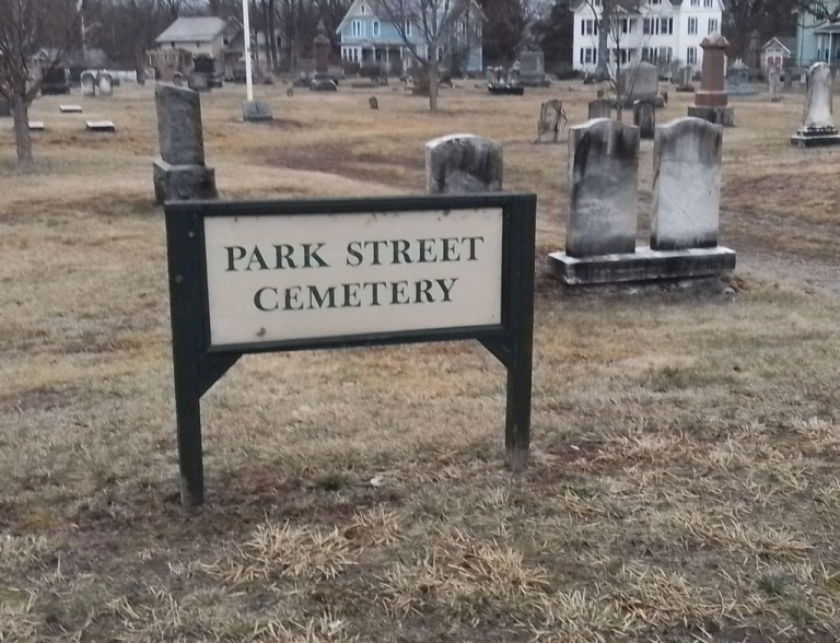 A weathered wooden sign reading "Park Street Cemetery" stands at the entrance of a historic graveyard in Florence, Massachusetts, with rows of aged headstones stretching into the background under a soft light.