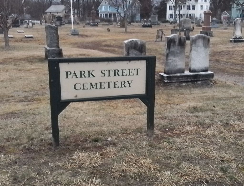 A weathered wooden sign reading "Park Street Cemetery" stands at the entrance of a historic graveyard in Florence, Massachusetts, with rows of aged headstones stretching into the background under a soft light.